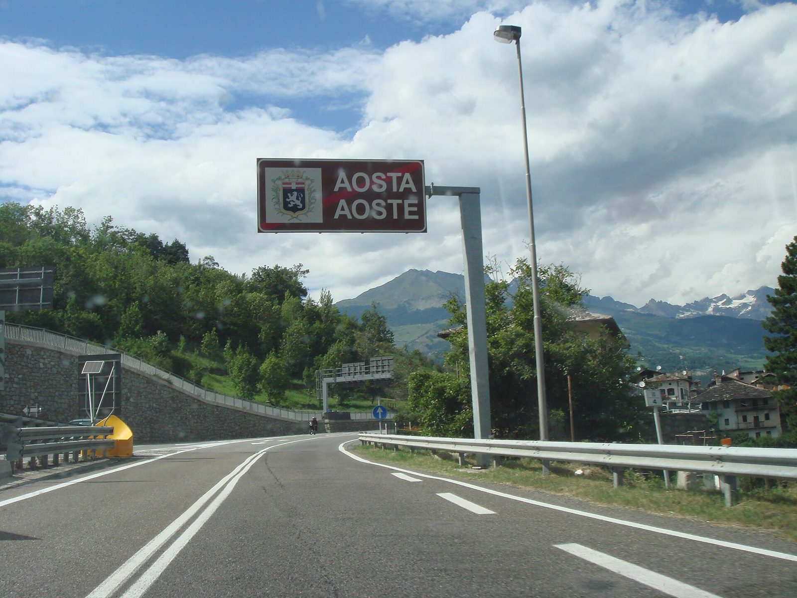 Cartello stradale bilingue con la scritta Aosta e Aoste e lo stemma della città, con le montagne sullo sfondo